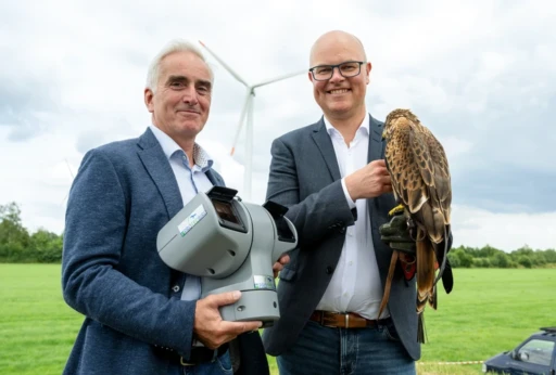Zwei lächelnde Männer in Jeans, Hemd und Blazer stehen auf einer Wiese vor einer Windturbine. Der linke hält ein graues, bewegliches Kamera-/Sensorsystem mit Protecbird-Logo, der rechte trägt einen Rotmilan auf einem Lederhandschuh. Im Hintergrund sind grünes Gras, Bäume und ein bewölkter Himmel zu sehen.