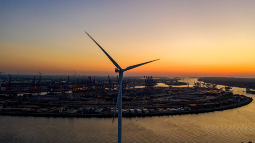 Blick aus der Vogelperspektive auf eine einzelne Windturbine am Ufer eines Hafens bei Sonnenuntergang: Die Rotorblätter drehen sich gegen den orange-violetten Himmel, unter ihnen spiegeln sich Hafenkräne und Containerbereiche im Wasser des Flusses.