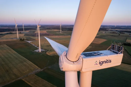 Vogelperspektive auf die Nabe und ein Rotorblatt einer weißen Windturbine mit der Aufschrift ‚ignitis‘ auf dem Maschinenhaus; im Hintergrund weite Ackerflächen und drei weitere Windturbinen unter klarem Himmel kurz nach Sonnenuntergang, das warme Abendlicht färbt die Landschaft in sanfte Töne.