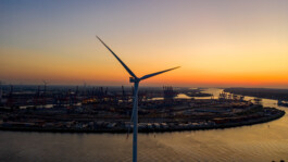 Blick aus der Vogelperspektive auf eine einzelne Windturbine am Ufer eines Hafens bei Sonnenuntergang: Die Rotorblätter drehen sich gegen den orange-violetten Himmel, unter ihnen spiegeln sich Hafenkräne und Containerbereiche im Wasser des Flusses. 
