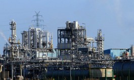 Petrochemische Anlage mit zahlreichen Rohrleitungen, Stahltürmen, Tanks und Verarbeitungsmodulen vor hellem Himmel; im Hintergrund ist ein hoher Strommast mit Hochspannungsleitungen zu sehen. 