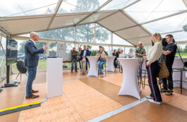 Ein Mann im Sakko und Jeans steht an einem weißen Stehpult und hält eine Präsentation in einem großen, transparenten Zelt. Vor ihm sitzen und stehen mehrere Zuhörer um hohe Stehtische, darunter eine Frau mit Kamera sowie ein Kameramann mit Videokamera. Durch die Zeltwände ist grünes Feld sichtbar.