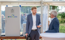 In einem Messezelt stehen zwei Männer in Hemd und Sakko vor einer Präsentationstheke mit Protecbird-Steuergehäusen; der linke Mann gestikuliert, während der rechte dem Gespräch lauscht und an einem weißen Stehtisch lehnt; durch die transparente Zeltwand ist Grünland zu sehen.