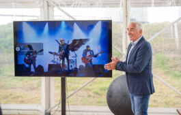 Thorsten Heinzen, ein Mann im Sakko und Jeans steht neben einem großen Flachbildschirm in einem transparenten Zelt. Auf dem Bildschirm ist eine Rockband mit drei Musikern zu sehen, die auf einer Bühne spielen und spotgeflutet werden. Im Hintergrund sind Wiese und Zeltkonstruktion erkennbar.