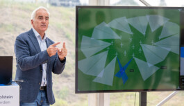 Thorsten Heinzen, ein Mann in dunklem Sakko und Jeans erklärt an einem großen Bildschirm in einem transparenten Zelt die Anzeige eines Windpark-Überwachungssystems: Auf dem Bildschirm ist auf grünem Hintergrund ein Kreis aus kleinen Windturbinen-Symbolen zu sehen, umgeben von grau-transparenten Sektoren und ein blaues Fadenkreuz in der unteren Mitte. Im Hintergrund durch die Zeltwand Wiese und Hügel.