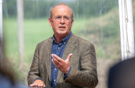 Ein älterer Mann mit dünner, runder Brille, grauem Haar und einem braunen Sakko über blauem Hemd steht in einem durchsichtigen Zelt vor einer grünen Wiese; er gestikuliert mit der rechten Hand, während er spricht und mit der linken Hand auf einem Stehpult ruht.