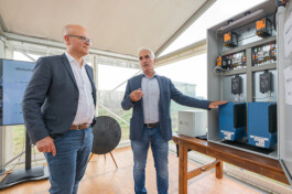 Zwei Männer in Jeans, Hemd und Sakko stehen in einem transparenten Messenzelt an einer Tischplatte mit offenem Steuergehäuse: Das Gehäuse zeigt oben orangefarbene Steuermodule und zahlreiche Kabel, unten zwei große blaue Elektronikgeräte; der grauhäutige Mann rechts deutet mit der Hand auf das Gehäuse, der Brillenträger links schaut aufmerksam zu.