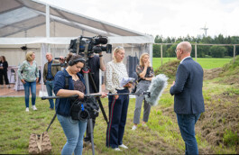 Ein Kamerateam filmt im Freien vor einem Zelt einen Mann im blauen Sakko: Eine Frau bedient das Mikrofon-Boom, ein Kameramann hält die Videokamera, zwei Journalistinnen mit Notizblock und Unterlagen stehen daneben, im Hintergrund grüne Wiese und Windturbine