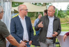 Zwei Männer im Gespräch vor einer Zeltwand und einer grünen Wiese: Links ein Glatzkopf mit Brille im dunklen Sakko und weißem Hemd, rechts ein Mann in grauer Jacke und hellblauem Hemd, der mit der rechten Hand gestikuliert.