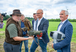 An einem Windpark-Feldrand steht eine Falknerin mit braunem Hut und Weste, die einen Rotmilan auf dem Handschuh hält, während ein Mann im Sakko und Lederhandschuh den Vogel entgegennimmt. Zwei weitere Herren im Hintergrund blicken lächelnd zu, einer hält eine Protecbird-Kamera. Im Hintergrund weite grüne Wiese, Windturbinen und bewölkter Himmel.