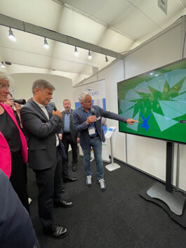 In einem Ausstellungszelt stehen mehrere Personen um einen großen Monitor: Ein Mann in Jeans, Sneakern und Sakko hält ein Mikrofon und zeigt auf auf dem Bildschirm dargestellte Windturbinen-Symbole und radarartige Sektoren; neben ihm steht ein Mann im dunklen Anzug ebenfalls mit Mikrofon in der Hand, zu ihrer Linken eine Frau im pinken Blazer. Im Hintergrund ist ein Banner mit ‚PROTECBIRD‘-Logo zu sehen.