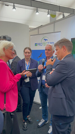 Vier Personen im Gespräch auf einem Messestand vor einem Banner mit ‚PROTECBIRD‘-Logo: Eine Frau im pinken Blazer gestikuliert links, daneben eine junge Frau mit Tablet in dunkelblauem Jackett, rechts stehen zwei Männer – einer im Jeans-Sakko hält ein Mikrofon, der andere im Anzug spricht in sein Mikrofon.