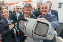 Auf einer Messe präsentiert eine junge Frau in dunkelblauem Blazer zwei Mikrofone, daneben stehen zwei Männer in Anzügen – einer hält lächelnd ein graues, drehbares Kamera-/Sensorsystem in der Hand (Thorsten Heinzen), der andere blickt neugierig darauf (Robert Habeck); im Hintergrund sind Messestände und weitere Besucher unscharf zu erkennen.“ 