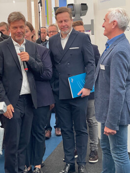 Mehrere Personen im Anzug stehen an einem Messestand: Ein Mann ganz links hält ein Mikrofon und spricht, daneben ein weiterer Mann im dunklen Doppelreiher-Anzug mit blauem Ordner in der Hand, rechts ein weiterer Herr im Jeans-Sakko lächelt. Im Hintergrund sind Ausstellungswände, Besucher und ein graues Kamerasystem zu sehen.