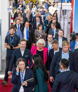 Ein dichtes Gedränge von Messebesuchern läuft durch einen Ausstellungsbereich auf rotem Teppich: Ganz vorne eine Frau im dunklen Blazer spricht ins Handy, neben ihr eine Frau mit langem dunklem Haar; dahinter mehrere Geschäftsleute in Anzügen und Blazern, unter ihnen eine Frau im pinken Blazer und mehrere Männer im dunklen Sakko; im Hintergrund sind Werbebanner, Kameraleute und weitere Besucher zu sehen.
