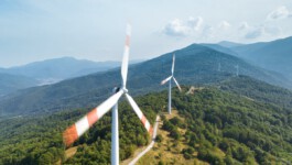 Drohnenaufnahme eines windreichen Höhenrückens: Im Vordergrund dreht sich ein Rotorblatt einer Windturbine mit rot-weißer Markierung, dahinter steht eine zweite Turbine auf einer schmalen Straße, gesäumt von grünen Wäldern und Hügeln unter hellem Himmel. 