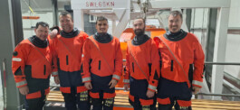 Fünf Männer stehen nebeneinander in einer Werkhalle und tragen leuchtend orangefarbene Überlebensanzüge mit reflektierenden Ärmel- und Hosensäumen. Sie lächeln in die Kamera, im Hintergrund ist ein Metallgerüst mit Beschriftung ‚SWL65KN‘ und Ausrüstungsteilen zu sehen.“ 