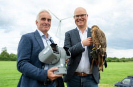 Zwei lächelnde Männer in Jeans, Hemd und Blazer stehen auf einer Wiese vor einer Windturbine. Der linke hält ein graues, bewegliches Kamera-/Sensorsystem mit Protecbird-Logo, der rechte trägt einen Rotmilan auf einem Lederhandschuh. Im Hintergrund sind grünes Gras, Bäume und ein bewölkter Himmel zu sehen. 