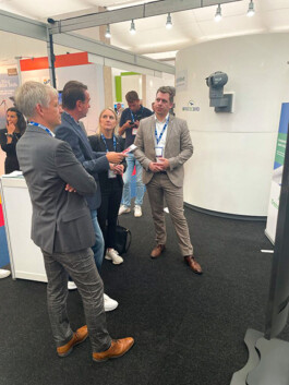 Vier Personen im Gespräch an einem Messestand: Drei Männer in Anzügen und eine Frau in schwarzem Blazer stehen vor einer runden, weißen Ausstellungswand mit einem montierten Protecbird-Kamerasystem; einer der Männer hält ein Faltblatt, alle blicken aufmerksam auf das Kameragerät.
