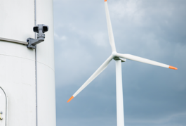 Nahaufnahme einer Windkraftanlage bei bewölktem Himmel: Links am Turmschaft ist ein graues, drehbares Kamera-/Sensorsystem montiert, rechts ragt ein weißes Rotorblatt mit orangefarbener Spitze ins Bild. 