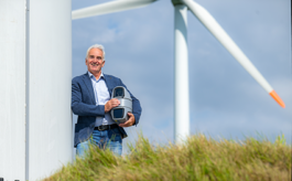 Geschäftsführer Thorsten Heinten, ein lächelnder Mann in Jeans, weißem Hemd und dunklem Sakko lehnt entspannt an der Basis einer Windkraftanlage und hält ein graues, drehbares Kamera-/Sensorsystem in beiden Händen; im Hintergrund ragt ein Rotorblatt der Windturbine in den bewölkten Himmel, im Vordergrund ist leicht verschwommenes Gras zu erkennen. 