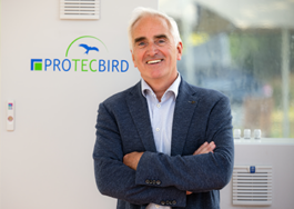 Geschätsführer Thorsten Heinzen: Ein lächelnder Mann mit grauen Haaren steht vor einer Wand mit dem Schriftzug ‚PROTECBIRD‘ und einem stilisierten Vogel-Logo; er trägt ein helles Hemd unter einem dunklen Blazer und hat die Arme vor der Brust verschränkt. Im unscharfen Hintergrund ist eine Glasfront oder ein Container erkennbar.“ 