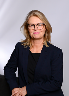 Porträt von Viola Heinzen mit schulterlangen, blonden Haaren und schwarzer, rechteckiger Brille; sie trägt ein dunkles Sakko und ein schwarzes Oberteil, lehnt mit einer Hand auf einer Rückenlehne ab und schaut selbstbewusst in die Kamera vor einem hellen, neutralen Hintergrund 