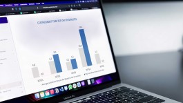Nahaufnahme eines MacBook Pro, auf dessen Bildschirm ein Balkendiagramm mit dem Titel „Curtailment time per day in minutes“ zu sehen ist. Die grauen Balken stellen die durchschnittliche tägliche Abregelungszeit der letzten 12 Monate dar, die blauen Balken die Abregelungszeit des aktuellen Tages für fünf Windturbinen (WTG1–WTG5): WTG1: 4,8 Min. (grau) vs. 0,0 Min. (blau) WTG2: 6,2 Min. vs. 9,0 Min. WTG3: 4,2 Min. vs. 5,2 Min. WTG4: 10,0 Min. vs. 19,6 Min. WTG5: 5,5 Min. vs. 0,0 Min. Am unteren Bildschirmrand ist die Mac-Menüleiste mit App-Icons und der Schriftzug „MacBook Pro“ auf der Gehäusekante zu erkennen. 