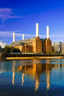 Großes, historisch anmutendes Backsteingebäude mit vier schlanken, weißen Schornsteinen am Ufer eines Flusses, dessen ruhige Wasseroberfläche das Kraftwerksgebäude und die Schornsteine spiegelt; im Hintergrund moderne Wohngebäude unter klarem, blauem Himmel. 