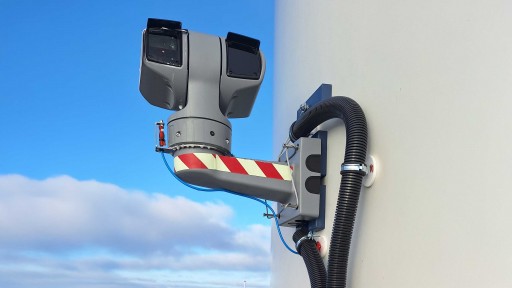 An einem hellen Turm einer Windkraftanlage montierte graue Halterung mit einem darauf sitzenden drehbaren Sensor- oder Kamerakopf mit zwei rechteckigen Gehäuseöffnungen; die Halterung trägt rot-weiße Warnstreifen, an der Wand verlaufen zwei schwarze Kabelschutzrohre, und ein dünnes blaues Kabel ist sichtbar; im Hintergrund blauer Himmel mit einzelnen Wolken.