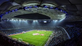 Panoramablick ins Inner eines fast voll besetzten Fußballstadions bei Abendlicht: Ein grüner Rasen mit Markierungen und Werbebannern, rundum Tribünen mit Zuschauerreihen; darüber hängt eine große, ovale Dachkonstruktion mit blauen und weißen Scheinwerfern, die das Spielfeld ausleuchten. 
