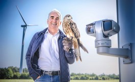 Geschäftsführer Thorsten Heinzen lächelnd in weißem Hemd und blauer Steppjacke steht vor einer Windkraftanlage auf einer Wiese und hält auf seiner ledernen Falke-Handschuh-Hand einen Rotmilan; rechts neben ihm ist an der Turmwand ein graues, drehbares Kamera-/Sensorsystem mit dem Schriftzug ‚PROTECBIRD‘ montiert. Im Hintergrund sind Bäume und blauer Himmel zu sehen. 