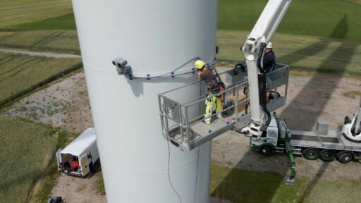 Zwei Arbeiter installieren mit einer Hebebühne eine Kamera am Turm einer Windkraftanlage.