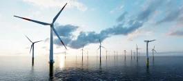 Offshore-Windpark auf dem Meer bei bewölktem Himmel und tiefstehender Sonne – zahlreiche Windkraftanlagen mit rot-weißen Rotorblättern stehen in einer Reihe im Wasser, die Fundamente gelb markiert. 