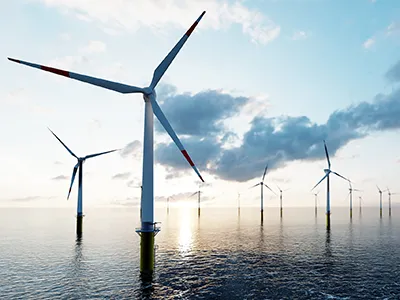 Offshore-Windpark auf dem Meer bei bewölktem Himmel und tiefstehender Sonne – zahlreiche Windkraftanlagen mit rot-weißen Rotorblättern stehen in einer Reihe im Wasser, die Fundamente gelb markiert.